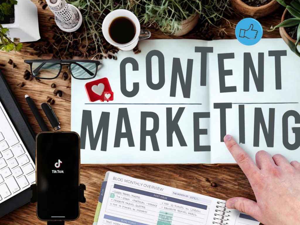 Schild mit der Aufschrift "Content Marketing", auf das eine Hand zeigt. darum eine Kaffeetasse, ein Laptop, ein Smartphone und ein Terminplaner