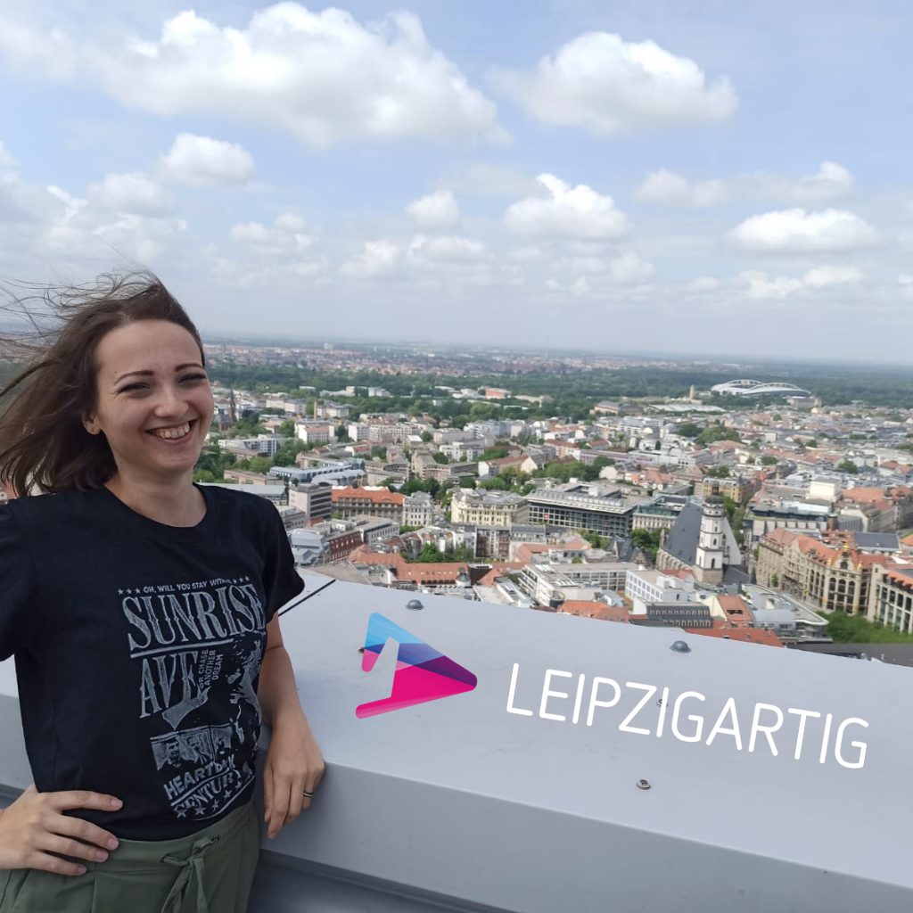 Jessica Jirschik auf dem MDR TOWER sieht lachend in die Kamera, hinter ihr sieht man die Dächer Leipzigs und das Logo für Leipzigartig
