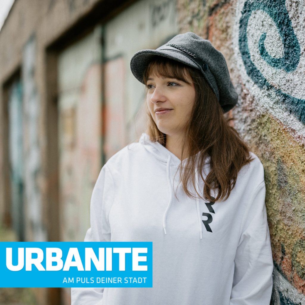 Jessica Jirschik lehnt an Graffiti-Wand in Leipzig und vorne der Schriftzug Urbanite - Am Puls deiner Stadt