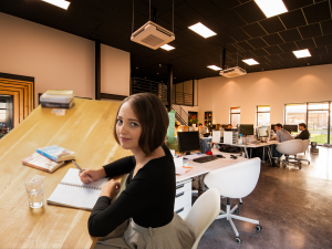 Jessica Jirschik sitzt am Schreibtisch vor Notizblock mit Stift in der Hand. Im Hintergrund ist ein Großraumbüro mit einigen Mitarbeitern zu sehen. Das Bild greift clever die Frage auf: Lohnt sich ein Freelancer oder SEO-Agentur eher zur Beauftragung